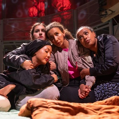 Kuvaelma näytelmästä. Neljä naista istuu vuodevaatteiden peittämällä lattialla toisiinsa nojaten. Näyttämön taustalla punasävyinen kukallinen verho. Kuva: Kansallisteatteri