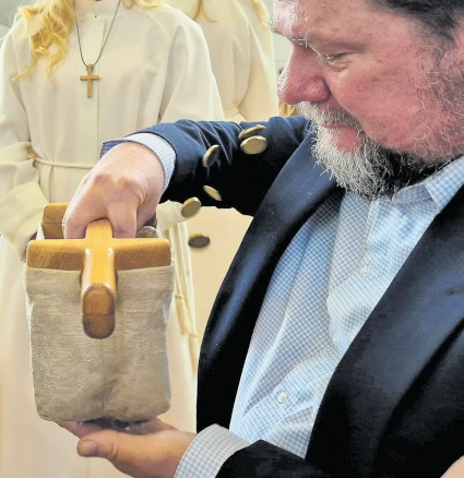 Mies napauttaa kädellään kolehtihaavin pohjaa ja kolikot pomppaavat ulos. Taustalla albaan pukeutuneita nuoria. Kuva: Saara-Sofia Järvinen