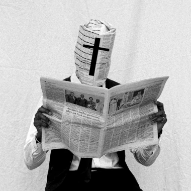 Mustavalkoinen kuva miehestä lukemasta sanomalehteä. Miehen pää on kääritty kokonaisuudessaan sanomalehteen, jossa kasvojen kohdalle on painettu risti. Olumide Adekunle / Unsplash