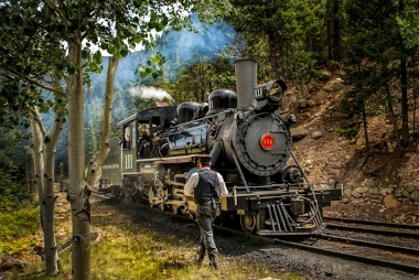 Kuvassa höyryveturi puiden reunustamassa kanjonissa. Veturimies kävelee kohti veturia. Kuva: crichmond / unsplash.com